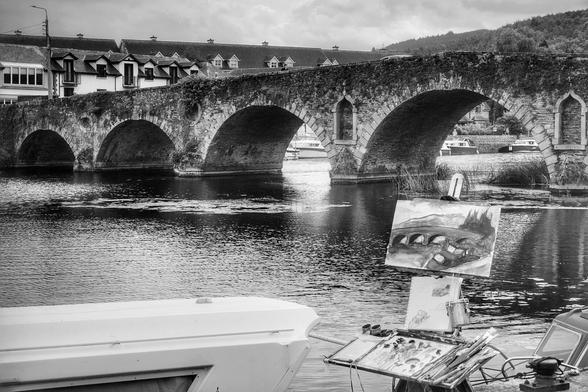 A black and white photo showing an artist's easel and painting kit in the foreground, with an arched stone bridge spanning the River Barrow in the background. The bridge is surrounded by buildings and lush hills under a cloudy sky.