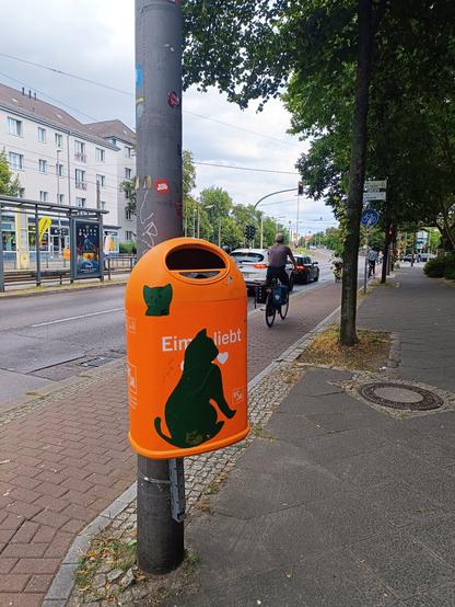 Das Bild zeigt einen orangefarbenen Mülleimer der Berliner Stadtreinigung (BSR) mit einem Aufkleber, der eine schwarze Katze darstellt. Der Aufkleber ist Teil der Kampagne „Eim liebt BSR“, die von der BSR ins Leben gerufen wurde, um für mehr Sauberkeit in Berlin zu werben.