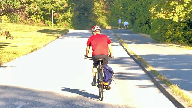 A cyclist on a two-lane paved road that is bordering by grass, trees and other vegetation.