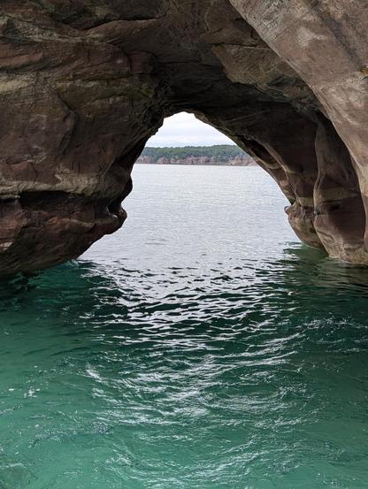 A rock formation on Grand Island, MI as viewed from a boat in Lake Superior. The water is a distinctive gitchi-gumi teal color with minimal waves, mostly from the wake of the boat. The beautiful sedimentary rocks form a triangular archway. You can see some more of the pictured rocks national shoreline through the other side under cloudy but still bright skies.