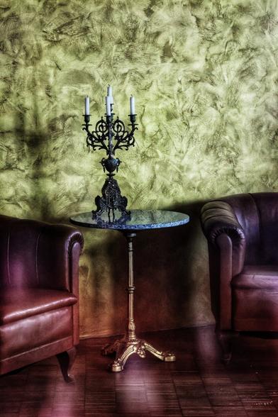A dimly lit, moody, close-up shot of a small, antique-style sitting area. A round, marble-topped pedestal table sits between two plush, brown leather armchairs. On the table, a dark metal candelabra with four white candles is the centrepiece. The background is a textured, mottled green and yellow wall. Light from an unseen source casts a shadow of the candelabra on the wall. The armchairs and the bottom of the image have a reddish-purple hue.