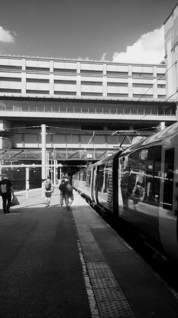 Black and white perspective shot looking along the side of a long line of railway carriages as they and the platform carry on out from under the shade of the overhead canopy into brilliant sunlight