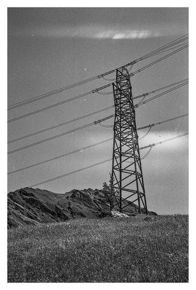 Black-and-white photograph of a power pole standing on the Tschinglenalp in the Swiss mountains. The pole is photographed from below. In the foreground, you can see some meadow, and in the background, a small rock massif rises toward the sky. In the sky itself, you can see a few wisps of cloud and a film defect or slight light incidence, which lends the mountain photo additional mystique.