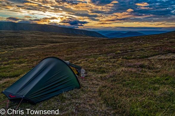 A small green tent pitched on a grassy slope in the hills under a colourful sky at dusk.