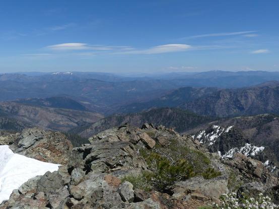 At the top of El Capitan.
Rocky peak as center foreground. A ridge falls away in steps to the left, a snow patch reaching toward its top to more left. The rest of the land drops off quickly to a rolling landscape with cliffs and gentler hills for layer after layer after layer. The sky has lumpy clouds echoing below. (They're lenticular clouds.)