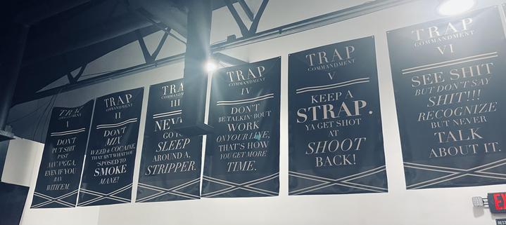 The image shows a series of six dark blue posters with white text, arranged horizontally on a wall. Each poster features a different commandment, labeled from I to VI, with a bold "TRAP COMMANDMENT" at the top. The text on the posters includes phrases such as "DON'T PUT SHIT IN YOUR MOUTH EVEN IF YOU WANT TO," "NEVER SLEEP AROUND A STRIPPER," and "KEEP A STRAP. YA GET SHOT AT SHOOT BACK!" The posters are evenly spaced and have a consistent design, with a white border and a decorative pattern at the bottom. The background is a plain white wall, and the lighting is bright, highlighting the posters. An exit sign is visible in the bottom right corner, indicating the location is likely a public or commercial space.