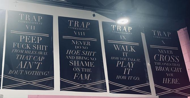 The image shows four large banners hanging on a wall, each with a dark blue background and white text. The banners are titled "TRAP COMMANDMENT" followed by Roman numerals VII, VIII, IX, and X. Each banner contains a distinct phrase in bold, capitalized white letters. The phrases are:"PEEP FUCK SHIT FROM MILES AWAY THAT CAP AIN'T BOUT NOTHING!""NEVER DO NO HOE SHIT AND BRING NO SHAME ON THE FAM.""WALK HOW YOU TALK IT, PLAY IT HOW IT GO.""NEVER CROSS THE ONES THAT BROUGHT YOU HERE."

The banners are evenly spaced and aligned, with a spotlight illuminating the top of the wall. The overall aesthetic is modern and urban, with a focus on bold, impactful statements.