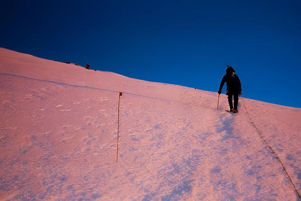 A mountaineer, roped in for safety, climbs up the snowy slope of Mt. Rainier at sunrise. The snow is illuminated with a reddish glow from the early morning light, contrasting sharply with the deep blue sky above. A flagged pole marks the route across the incline, and faint climbers can be seen further up the ridge. The scene captures the stark beauty and solitude of high-altitude alpine climbing.