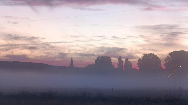 Morgenhimmel nach unten zurück, die Sonne ist noch nicht aufgegangen. 
Am Horizont Silhouetten von Bäumen und einer Kirche. Davor Bodennebel und Wiesen.