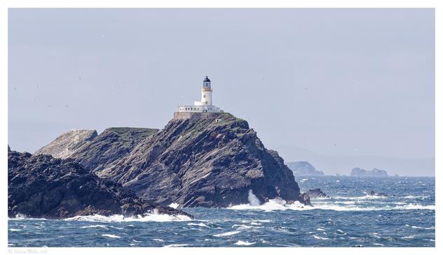 A lighthouse stands atop a rocky cliff, surrounded by turbulent ocean waves. The sky is partly cloudy, with distant land visible in the background. Seabirds are flying around the lighthouse, adding to the coastal scene.