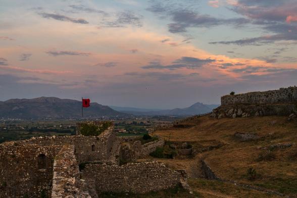 View of a ruined fortress with the Albanian flag waving, mountains and fields in the distance at dusk.