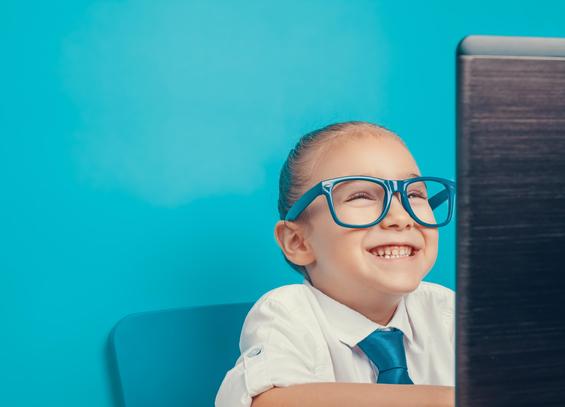 Ein Kind mit übergroßer farbiger Brille sitzt in Hemd und Krawatte grinsend vor einem Bildschirm. Foto: Myst · Adobe Stock
