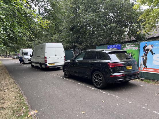Bloated black Audi SUV reg Y11XVX and two white vans completely blocking the pedestrian half of North Meadow Walk, forcing people into the adjacent cycle lane. There are festival hoardings and trees behind.