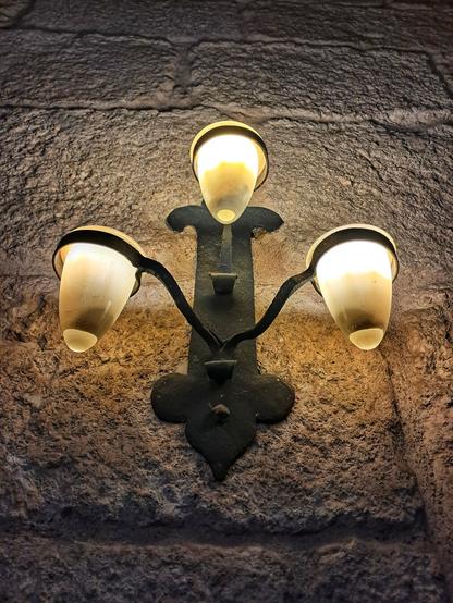 [pt] candeeiro em ferro com três lâmpadas, numa parede de pedra antiga

[en] iron lamp with three bulbs, on an old stone wall