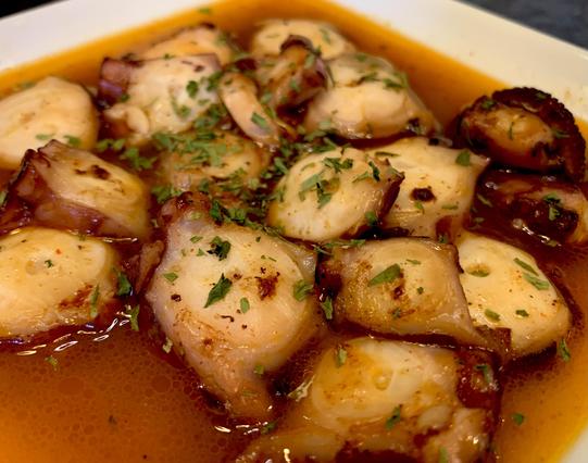Sautéed sliced octopus in a red-brown sauce, sprinkled with dried parsley and served on a white square plate.