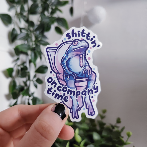 Hand holding up a sticker of a frog with a tie sitting on a toilet titled "shittin' on company time"