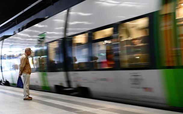 Auf dem Bahnsteig wartet ein in beigefarbener Hose, heller Mütze und hellbrauner Lederjacke gekleideter älterer Herr, die Arme hinter dem Rücken und eine blaue Tasche haltend, auf den just von rechts ins Bild hinein heranbrausenden, bewegungsunscharfen Zug, dessen Inneres zu verschwommen ist, als dass man Details ausmachen könnte.