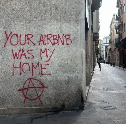 a graffiti on a wal: “YOUR AIRBNB WAS MY HOME” followed by the anarchism symbol