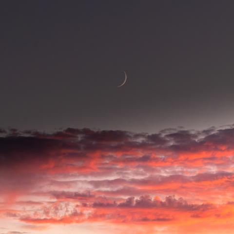 A thin crescent moon in the sky over some cloud catching the sunset rays.