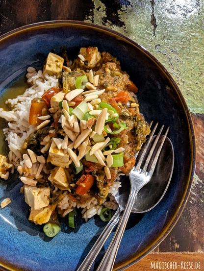 Dunkler Holztisch mit grünen Farblflecken. Darauf ein blauer, tiefer Teller. Darin Basmatireis, bedeckt mit einer dicken Sauce Gemüsecurry. Stellenweise sind weißliche Tofuwürfel und Paprikastücke erkennbar. Als Topping darübergetreut sind grüne Frühlingszwiebelringe und geröstete Mandeln.