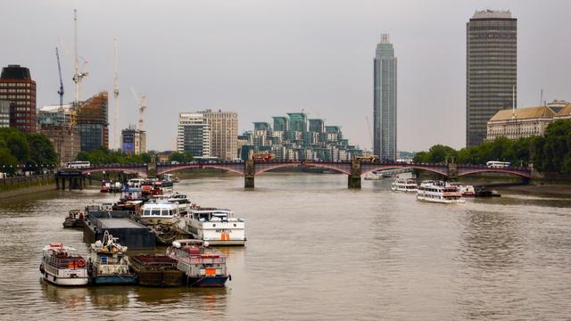Overcast sky above the River Thames featuring multiple boats moored and moving, with Lambeth Bridge in the background. Skyscrapers and construction cranes can also be seen, illustrating a cityscape likely in an urban setting.
