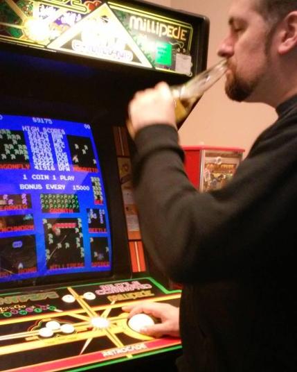 A guy drinking a beer and playing a Centipede/Millipede arcade cab