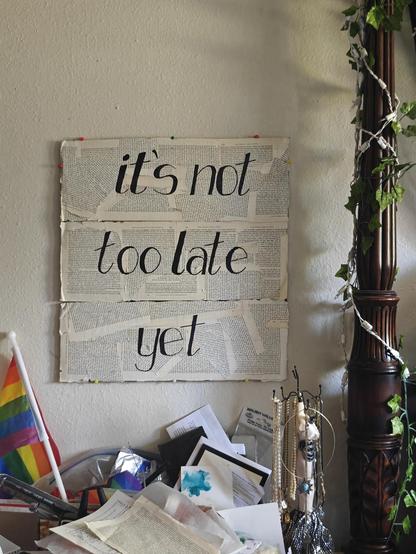 piece of handmade art hanging on a bedroom wall over a messy desk. main discernable item in the clutter is a small pride flag on a tiny pole. the art piece is three panels covered with old torn book pages, and emblazed with large black text, which reads:

its not too late yet