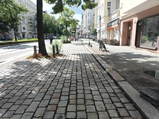 Endlose, 3m breite Kopfsteinpflasterschneise zwischen Bauminseln am Fahrbahnrand und Gehweg. Hier könnte (nach Beseitigung des Rumpelpflasters) problemlos ein Radweg verlaufen, aber es wird ( wenn nicht gerade wie zum Zeitpunkt des Fotos Bauarbeiten es verbieten) durch massenhaft quer stehende Stehzeuge mivbraucht. Radlys sollen stattdsssen hochgradig gefährdet im Rangierbereich links vom Stehblech fahren