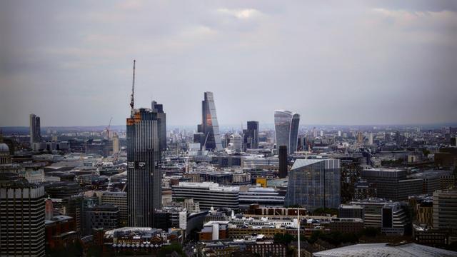 Panoramic view of a city skyline featuring a mix of modern skyscrapers of various shapes including cylindrical and angular designs, under a cloudy sky. The foreground shows densely packed buildings with a tower under construction towards the left.