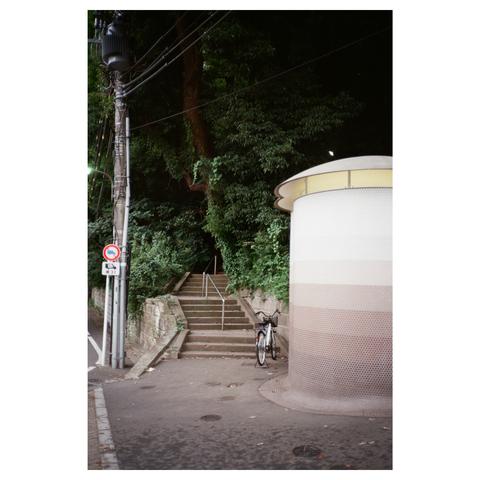 A Tokyo Toilet from the movie "Perfect Days". It's a cylindric shape to the right side of the image. Beside it is a parked bicycle, and behind it a forest, with a stone stair leading into it.