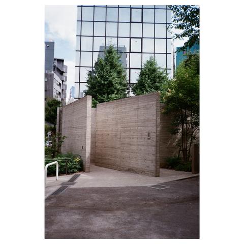The restroom building consists of two raw concrete walls, angled to each other, framed by some trees. In the background, a high-rise building with mirrored windows can be seen.