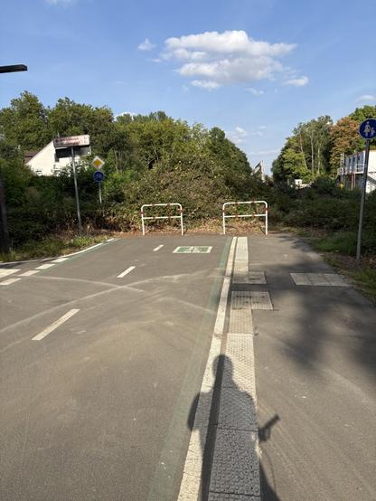 Das Bild zeigt eine Straße mit einem Sattelpunkt und einer Rampe. Der Hintergrund besteht aus Bäumen und einem blauen Himmel. Die Straße hat einige Markierungen und ein Schild, das auf einen Parkplatz hinweist. Ein Schatten einer Person ist auf einem Piktogramm zu sehen. Die Umgebung ist tagsüber, und die Straße sieht gepflegt aus.