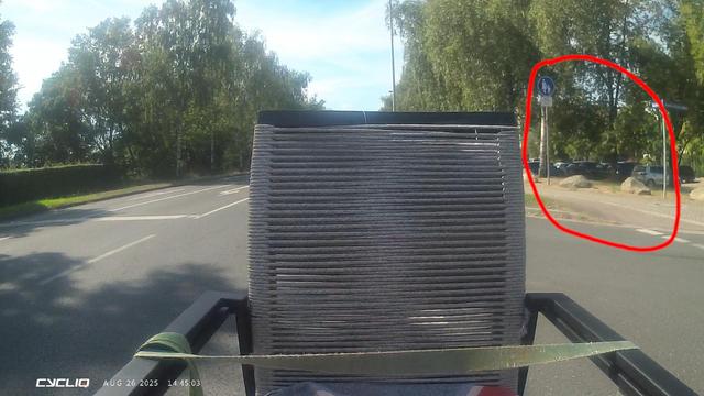Das Bild wurde von einer Fahrraddashcam aufgenommen und zeigt eine Straße, bei der rechts ein Gehweg mit halbseitiger roter Pflasterung zu sehen ist. Der Gehweg ist mit  Verkehrszeichen 239 "Gehweg" und "Radfahrer frei" Zusatz ausgeschildert. Auf dem Lastenrad, auf dem die Dashcam befestigt ist, wurden auf der vorderen Ladefläche zwei Stühle befestigt, daher ist ein Großteil des Bildes mit einem Stuhl verdeckt.