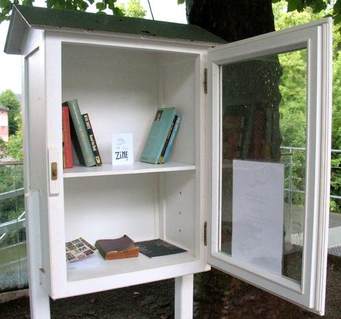 White little free library at the corner of a small square with trees in the background. The glass door is open. There are two shelves with each a few books and on the second shelf a mini-zine called Hoe maak je een zine (in Dutch).