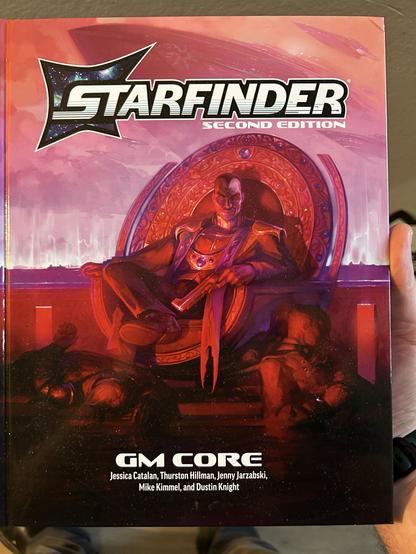 A large TTRPG book. At the top it reads “Starfinder Second Edition” and at the bottom “GM Core”. In the center in painterly tones of red, pink, and purple, sits a male presenting individual on a circular throne and holding a gun. At his feet and laying against the throne are two bodies, possibly his victims.