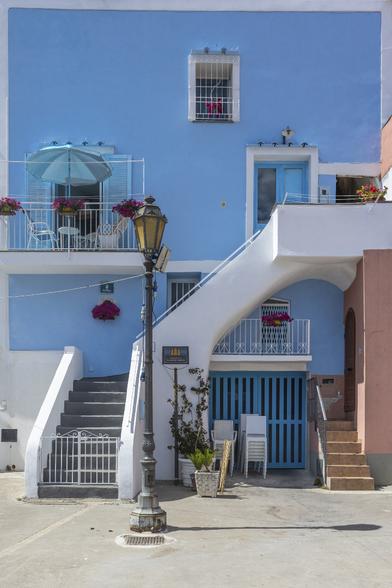 A colorful house of Procida island in a typical Mediterranean style rendered in plaster and painted in pastel blue.  An external plain white staircase crosses the facade in diagonal. Painted frames around simple windows add an other touch of white on the facade.
