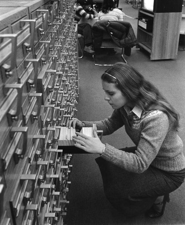 A black and white photo of a woman in front of a huge case of tiny drawers. She has one open and is searching through thousands of cards looking for specific books in the library likely by title, author or subject