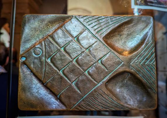 A rectangular, weathered bronze door handle features a stylised fish. The fish's body is made up of a grid of raised diamond shapes. Its round eye is a small circle with a raised dot in the centre. The fish has a triangular tail and two fins, all with incised lines. The bronze has a slight verdigris patina. The image is a close-up, and the door handle appears to be mounted on a glass door.
