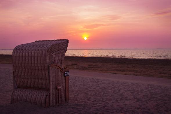 Sonnenuntergang an der Ostsee. Links im Vordergrund steht ein Strandkorb, den man halb seitlich von hinten sieht. Er blickt aufs Meer. Der Sandstrand zieht sich die untere Hälfte des Bildes. Die Sonne steht direkt über einem dichten Wolkenband am Horizont, direkt über ihr beleuchtet sie ein Stück Wolke. Der Himmel leuchtet in verschiedenen Orangetönen, zum Meer hin mit einem Hauch Lila.