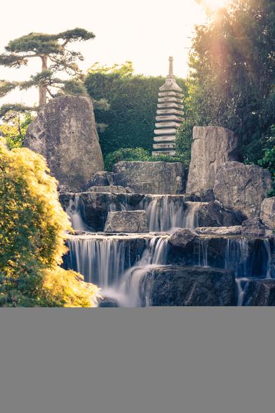 A serene Japanese-style garden scene with a multi-tiered stone pagoda standing among tall rocks and lush greenery. In the foreground, a gentle waterfall cascades down dark boulders into a small stream, with smooth white pebbles scattered at the base. Sunlight filters through the trees, creating a warm golden glow.