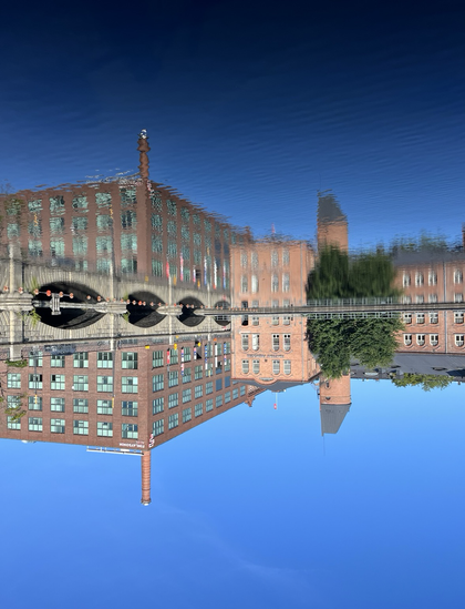 Ylösalaisin käännetty kuva Tampereen keskustasta. Alhaalla tyyni sininen taivas, keskellä punatiilisiä Finlaysonin tehdasrakennuksia, ylhäällä tyven, hieman väreilevä vesi symmetrisine heijastuksineen.