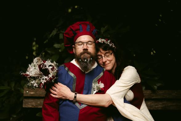This medieval-themed wedding photo by European Elopement and Wedding Photographer Sturmsucht shows a joyful couple in an intimate embrace outdoors. The groom wears a striking blue and red tunic with gold trim and a distinctive red medieval cap with blue accents. He has a full dark beard, glasses, and stands confidently in the center. The bride, wearing glasses and a delicate floral crown of white and red flowers, embraces him from behind in a flowing cream-colored dress with medieval-style sleeves. She holds a rustic bouquet of dried flowers and herbs in muted burgundy and white tones. Both wear wedding rings and beam with happiness. The setting features a beautifully blurred background of lush green foliage, creating a forest-like atmosphere with soft, warm lighting that enhances the romantic, timeless quality of their medieval celebration.