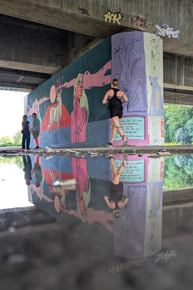 Das Bild zeigt eine großflächige Wandmalerei, die sich auf einem Betonpfeiler unter einer Brücke befindet. Die Szenerie wird in einer Pfütze gespiegelt. Diese Spiegelung verstärkt die visuelle Wirkung des Kunstwerks und der Personen im Bild. Die Wandmalerei ist abstrakt und surreal gestaltet. Im Zentrum steht zwei auffällige, pinkfarbene humanoide Figuren mit langen blonden Haaren. Dazwischen ist ein weißer Hirsch mit pinkem Geweih zu sehen, am Rand stilisierte pflanzliche Formen. Die Farbgebung ist kräftig und kontrastreich, wobei Pink als dominierende Farbe mehrfach betont wird. Am Rand befindet sich der Text „the bridge and the river are sacred thresholds to other worlds. you might see the old gods passing through but beware the watchers." Eine Joggerin und ein Händchen haltendes Paar sind vor der Malerei zu sehen.