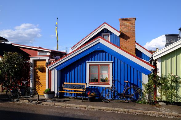 Eingeschossiges blau gestrichenes Holzhaus mit rot gestrichenen Fensterrahmen neben weiteren kleinen bunten Häusern. Vor dem Haus steht eine Bank und ein altes Fahrrad. Im Hintergrund steht ein Fahnenmast, von dem eine schwedische Fahne schlaff herunterhängt.
