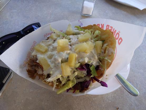 In einer weißen Papiertüte befindet sich ein üppig gefüllter Döner mit viel Salat, Fleisch und Joghurtsoße. Darüber verteilt sieht man viele kleine gelbe Ananaswürfel.