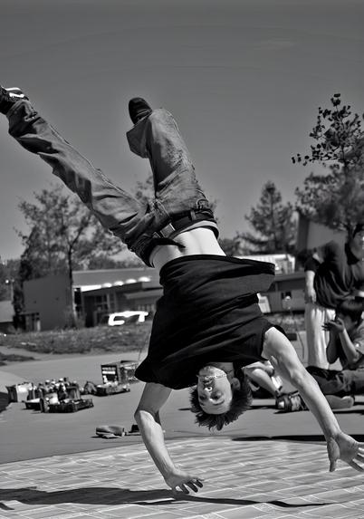 A young man performs a handstand in the air while wearing a black t-shirt and jeans, framed against a blurred background of a park and spectators.