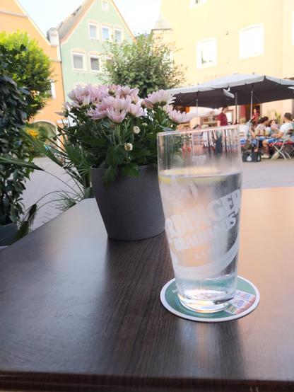 Ein Blick aus der Ich-Perspektive von einem Café-Tisch, auf dem ein hohes Glas mit Wasser, Eiswürfeln und einer Zitronenscheibe steht. Auf dem Glas ist der Schriftzug "ERDINGER BRAUHAUS" zu erkennen, und es steht auf einem runden, grün-weißen Untersetzer. Daneben befindet sich ein grauer Blumentopf mit vielen zartrosa Blüten, vermutlich Chrysanthemen. Im Hintergrund öffnet sich ein belebter Platz oder eine Gasse mit mehreren Personen, die entspannt an Tischen unter großen, dunklen Sonnenschirmen sitzen. Bunte, traditionelle Gebäude mit mehreren Fenstern säumen den Platz – eines ist hellgrün, ein anderes hellgelb oder orange. Die Szene wirkt sonnig und lädt zum Verweilen ein.