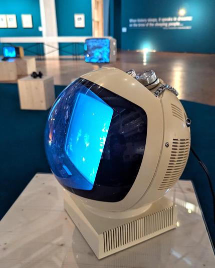 A vintage spherical television set with a cream-colored body and a large curved blue-tinted screen, showing abstract visuals. The TV is placed on a light wooden pedestal in a gallery with teal-colored walls, where other retro monitors and framed artworks are displayed in the background.