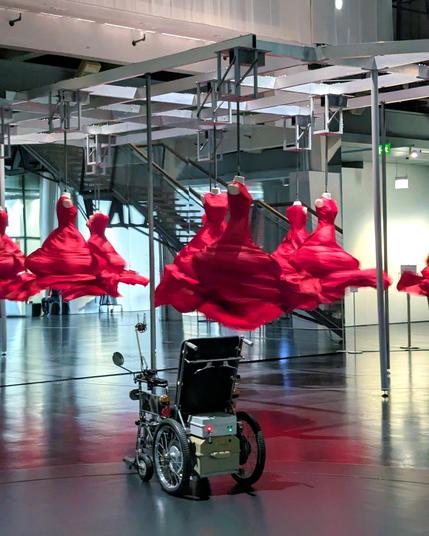 An art installation with several red fabric sculptures resembling spinning dresses, suspended from the ceiling in motion, creating a swirling effect. In front of them sits a modified electric wheelchair, placed as part of the exhibition. The space has reflective flooring and mirrors, enhancing the dynamic movement of the red forms.