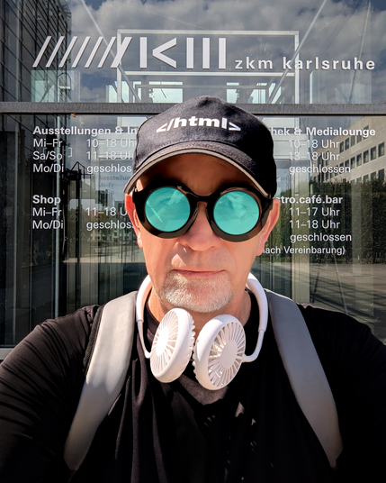 A man is taking a selfie in front of the entrance to ZKM Karlsruhe (Zentrum für Kunst und Medien). He is wearing a black baseball cap with white text </html>, round black sunglasses with bright turquoise reflective lenses, a white goatee, a black shirt, a light gray backpack, and a white portable neck fan hanging around his neck. Behind him is a glass door with white signage showing the opening hours in German: "///// ZKM zkm karlsruhe" at the top, large bold white letters. The background shows reflections of modern buildings and glass architecture in bright daylight.